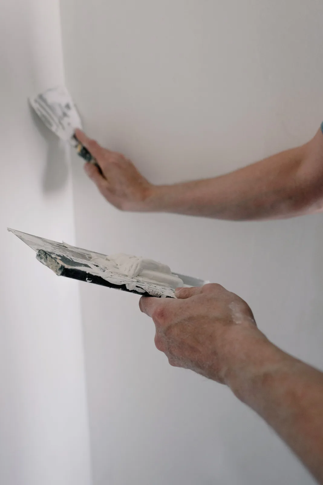 Two hands smoothing joint compound into a wall corner with taping knives