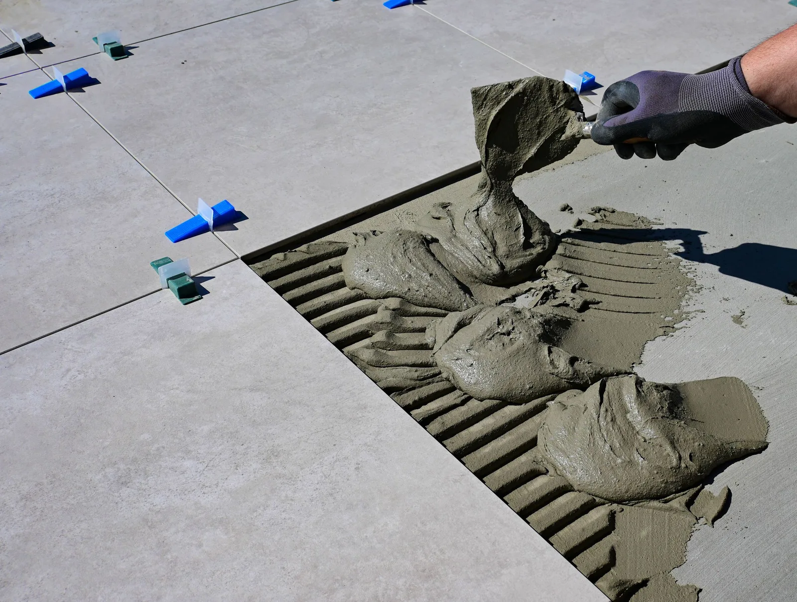 A gloved hand troweling mortar onto a floor between large tiles with leveling clips